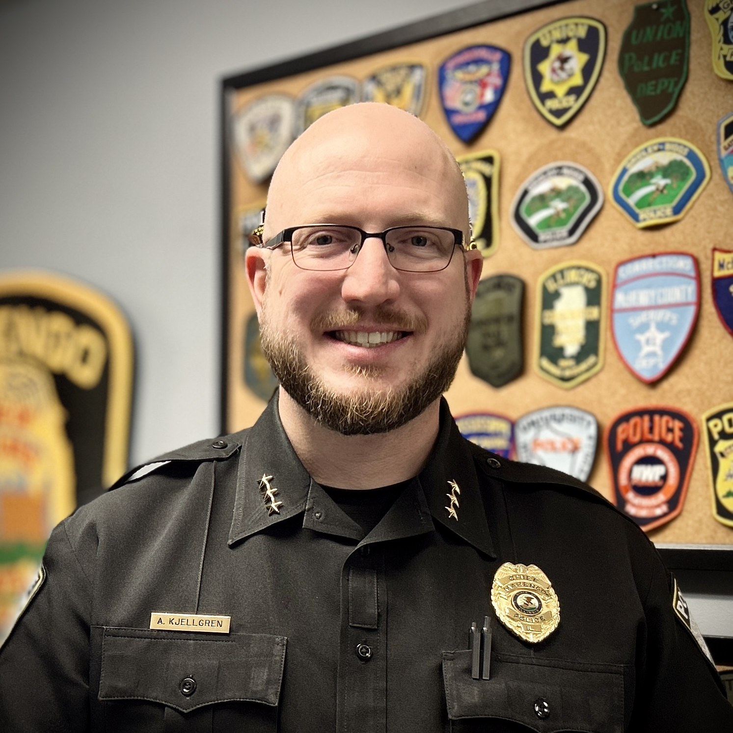 Marengo Police Chief Andrew Kjellgren, smiling, in a dark uniform in front of a board of patches from other police departments.